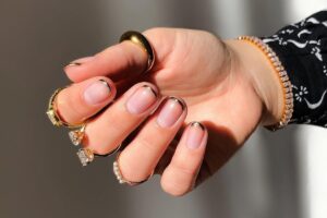 A imagem mostra uma mão com unhas bem cuidadas, decoradas com uma fina linha metálica na ponta, em um estilo conhecido como "francesinha invertida" ou "unhas francesas modernas". A pessoa está usando vários anéis dourados, alguns com pedras grandes e outros com detalhes de pérolas. O pulso tem uma pulseira brilhante. A luz na imagem destaca o brilho dos acessórios e o esmalte das unhas.