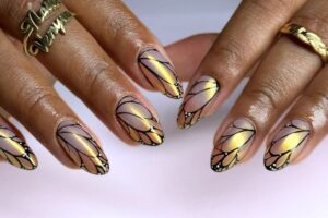 A imagem mostra mãos com unhas decoradas em formato de asas de borboleta. As unhas têm um design em tons de dourado e lilás, com contornos pretos e detalhes em branco nas pontas, imitando as delicadas asas de uma borboleta. Os dedos também exibem anéis dourados, complementando o visual elegante e artístico.