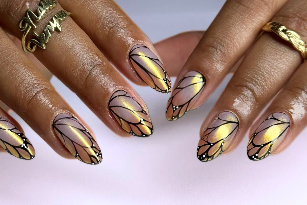 A imagem mostra mãos com unhas decoradas em formato de asas de borboleta. As unhas têm um design em tons de dourado e lilás, com contornos pretos e detalhes em branco nas pontas, imitando as delicadas asas de uma borboleta. Os dedos também exibem anéis dourados, complementando o visual elegante e artístico.