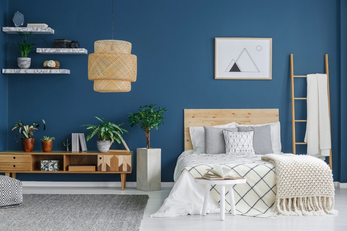 A imagem retrata um quarto moderno com parede azul, cama de madeira clara e colcha branca com detalhes geométricos. Prateleiras e plantas decoram o ambiente, enquanto uma luminária de palha e uma escada de madeira adicionam charme natural.