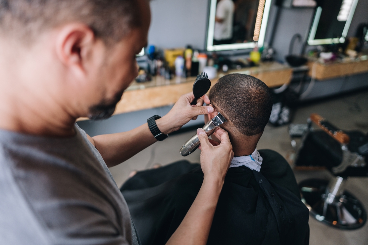 Imagem colorida de homem cortando cabelo de outro homem sentado em uma cadeira dentro de uma barbearia. Metrópoles