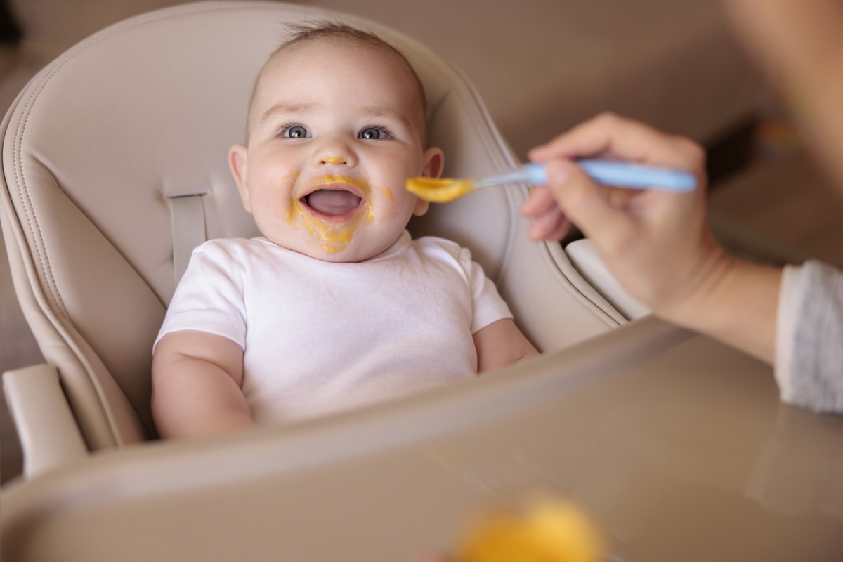 Mãe alimentando seu filho alegre enquanto apresentava a primeira refeição sólida, bebê todo bagunçado e com mingau sentado na cadeira alta e rindo. Metrópoles