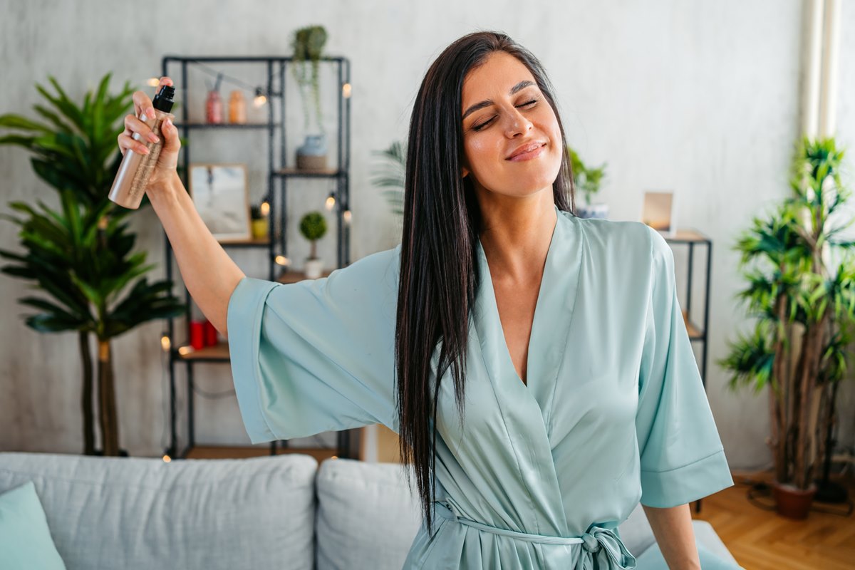 Foto colorida de mulher pulverizando spray no cabelo. Ela está em uma sala - Metrópoles