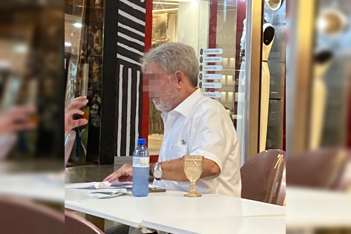 Imagem colorida de um homem de cabelo e barba brancos, vestido com camiseta branca, sentado em uma mesa branca com uma garrafa de plástico com água ao lado