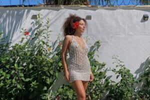 Julia Rodrigues usando vestido de crochê branco com plantas ao fundo e flor presa na orelha