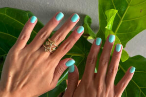 Mãos femininas com unhas curtas e arredondadas pintadas em um tom turquesa vibrante. Os dedos exibem anéis dourados delicados, e folhas verdes no fundo criam contraste natural, destacando a cor fresca e moderna inspirada nas tendências de esmaltes para 2025.