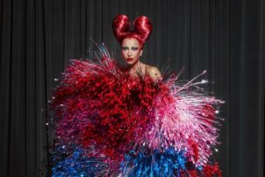 sabrina Sato em um traje extravagante com uma explosão de texturas e cores vibrantes em vermelho, rosa e azul. O cabelo vermelho em formato dramático complementa o visual, destacando-se contra um fundo preto elegante.