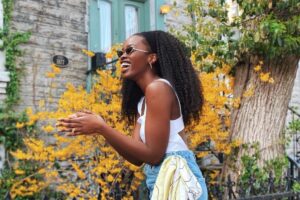 Mulher com cabelo afro longo e volumoso preso com faixa preta, usando óculos escuros, blusa branca e jeans, sorrindo em frente a um arbusto amarelo vibrante.