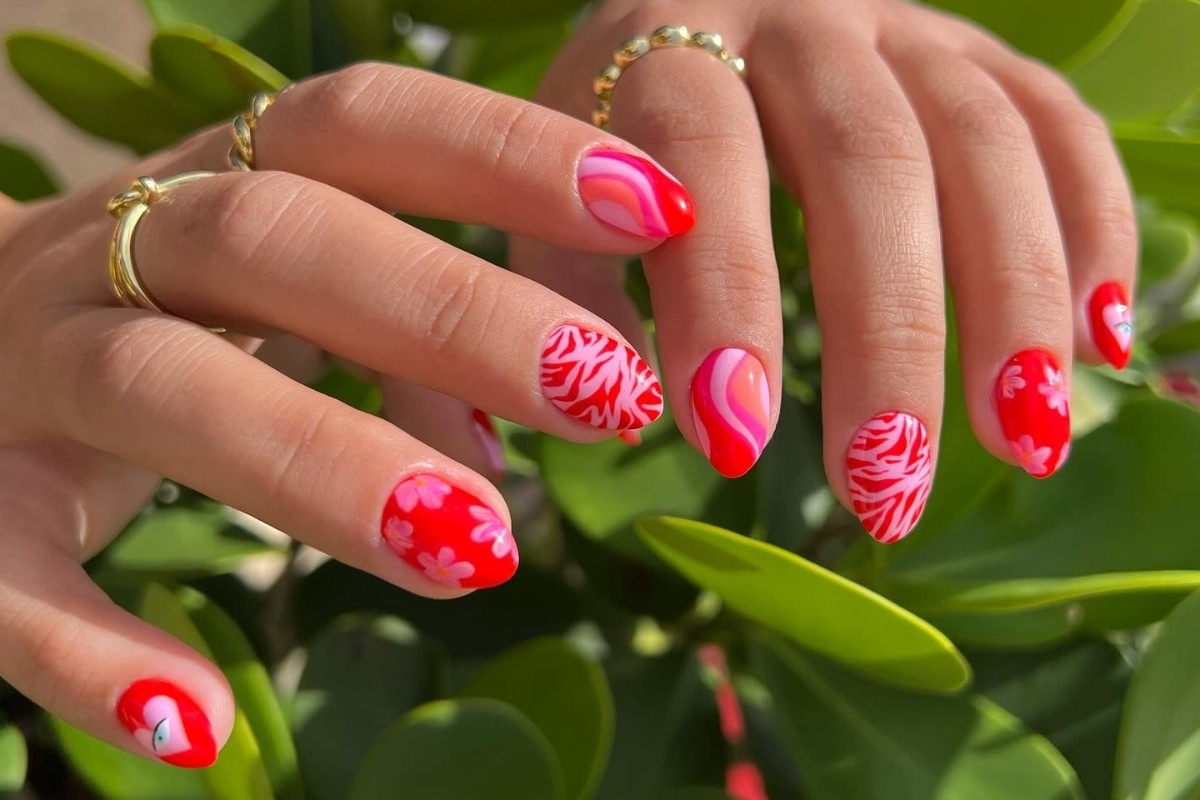 Mãos exibindo estilos de unhas curtas, decoradas com esmalte vermelho vibrante e designs variados, incluindo flores, padrões abstratos ondulados em rosa e branco, e estampas tropicais. Anéis dourados adornam os dedos. Fundo de folhas verdes complementa o visual criativo e moderno.